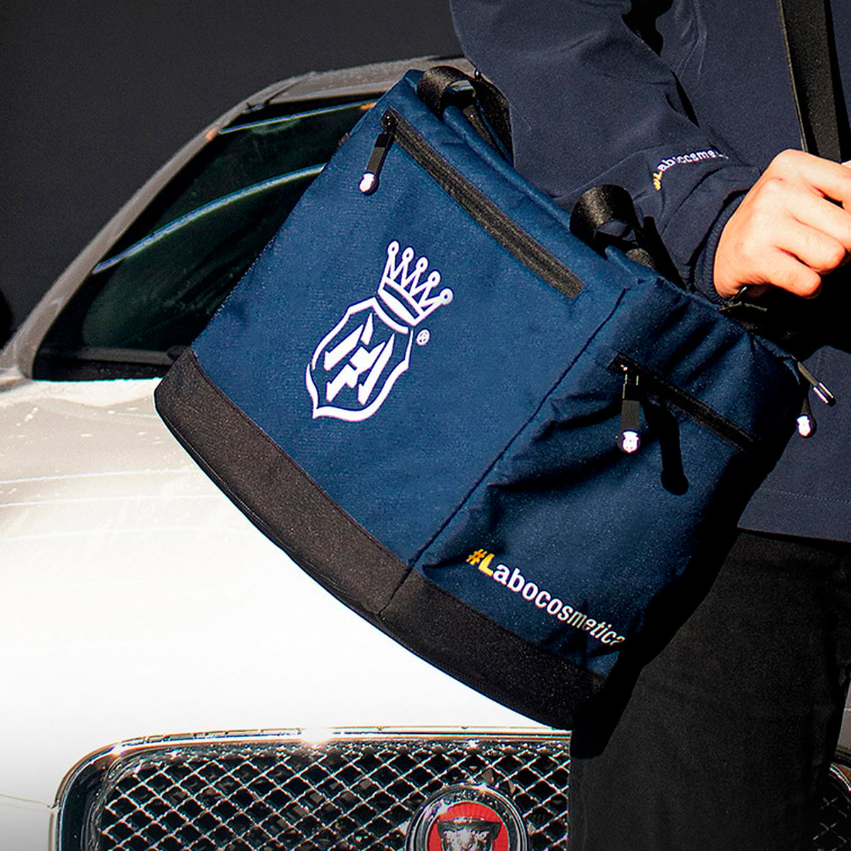 A navy blue tote bag with black trim and white branding, held by a person in front of a white car. The bag features a white crown-and-shield logo and the text 'labocosmética'.