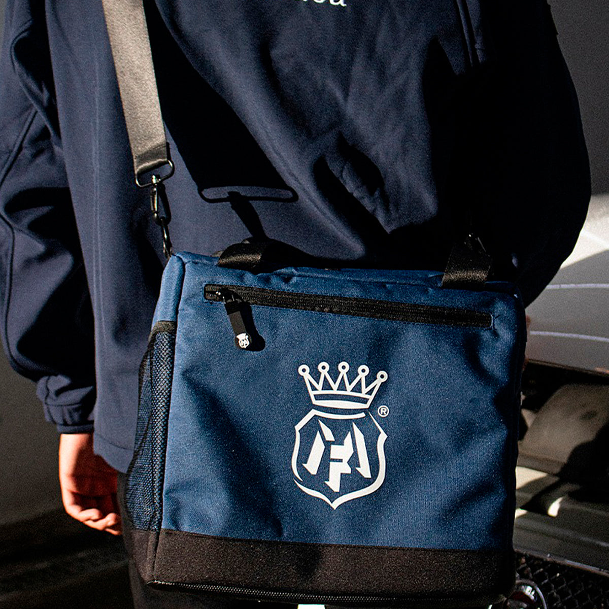 A navy blue crossbody bag with a white crown and monogram logo, worn by a person in a navy jacket.