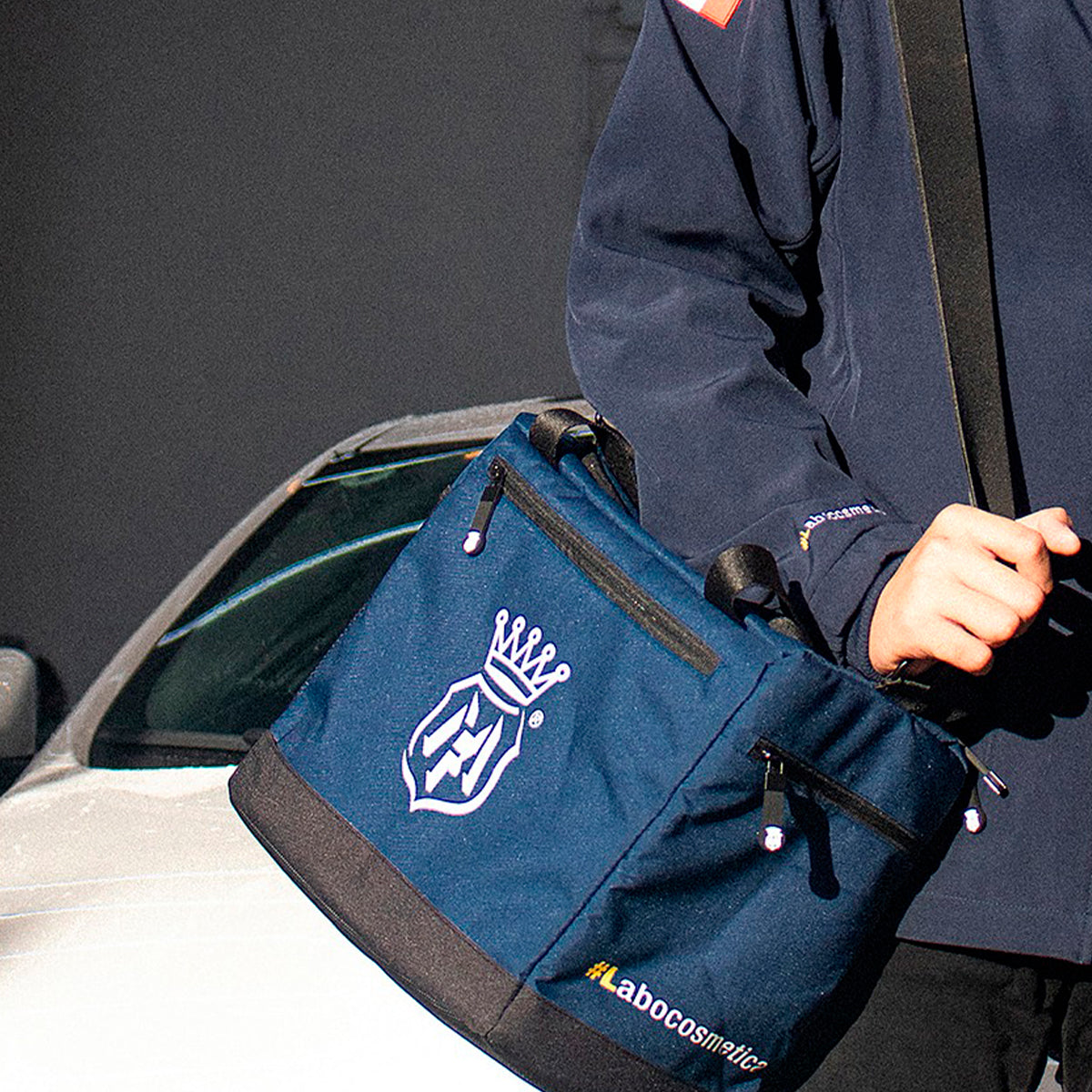 A navy blue tote bag with black trim and white logo graphics, carried by a person wearing a dark jacket next to a white car.