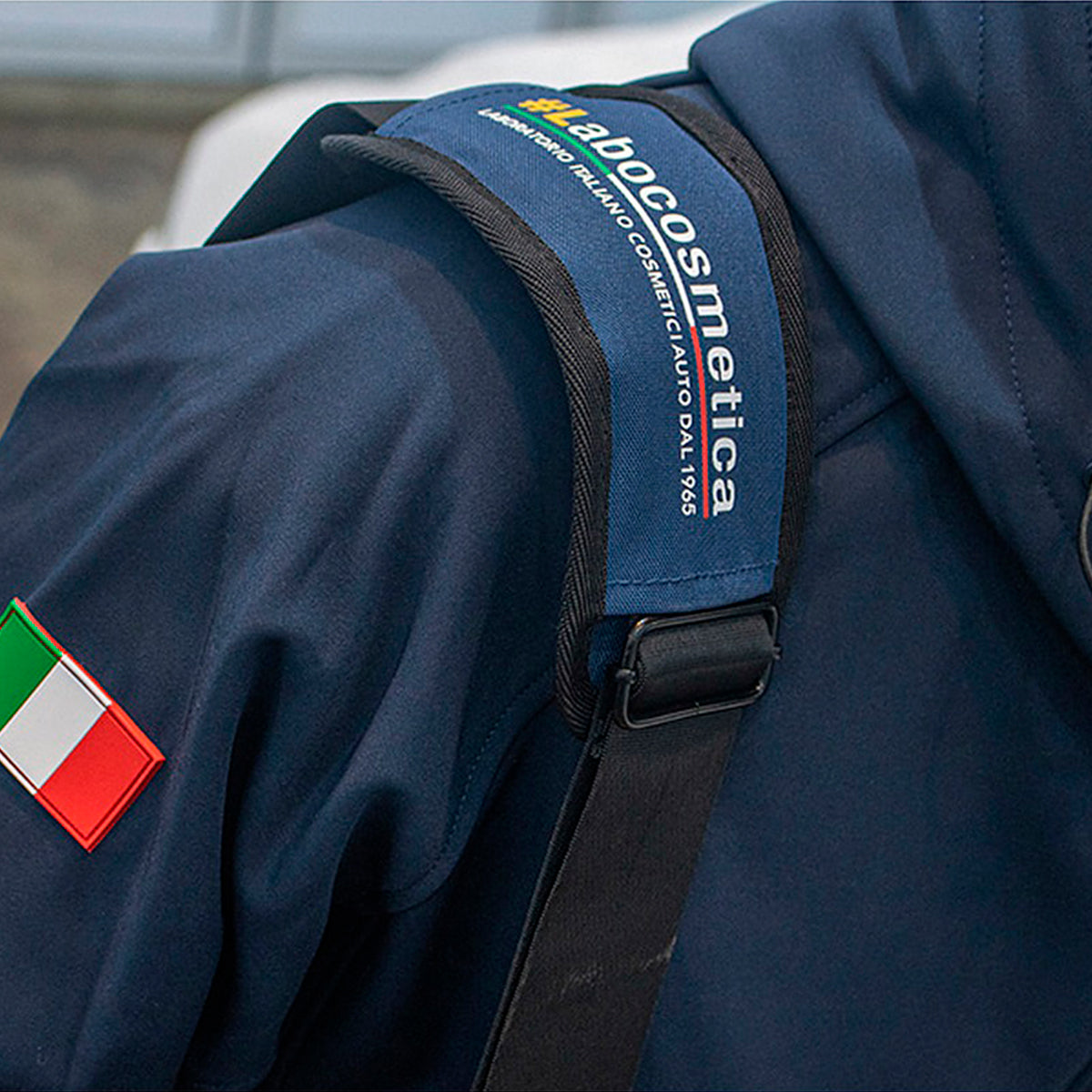 Close-up of a navy blue jacket or uniform with a black shoulder strap across the upper arm. The strap has a blue fabric label reading 'MocoCosmologica' with smaller white text.