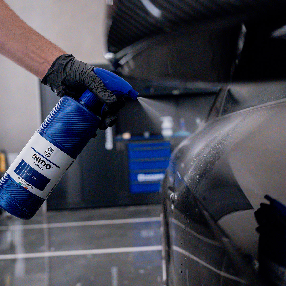 Person using a blue spray bottle labeled 'INITIO' on a car in a garage.