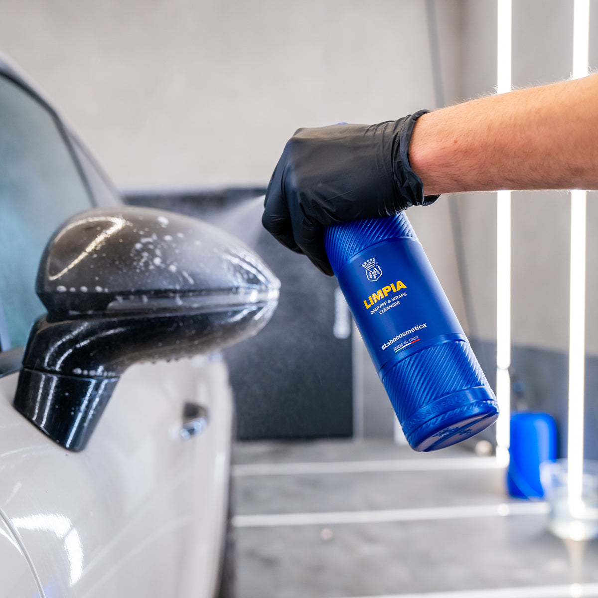 Person cleaning car mirror with a blue bottle labeled 'LIMPIA' in a garage setting.