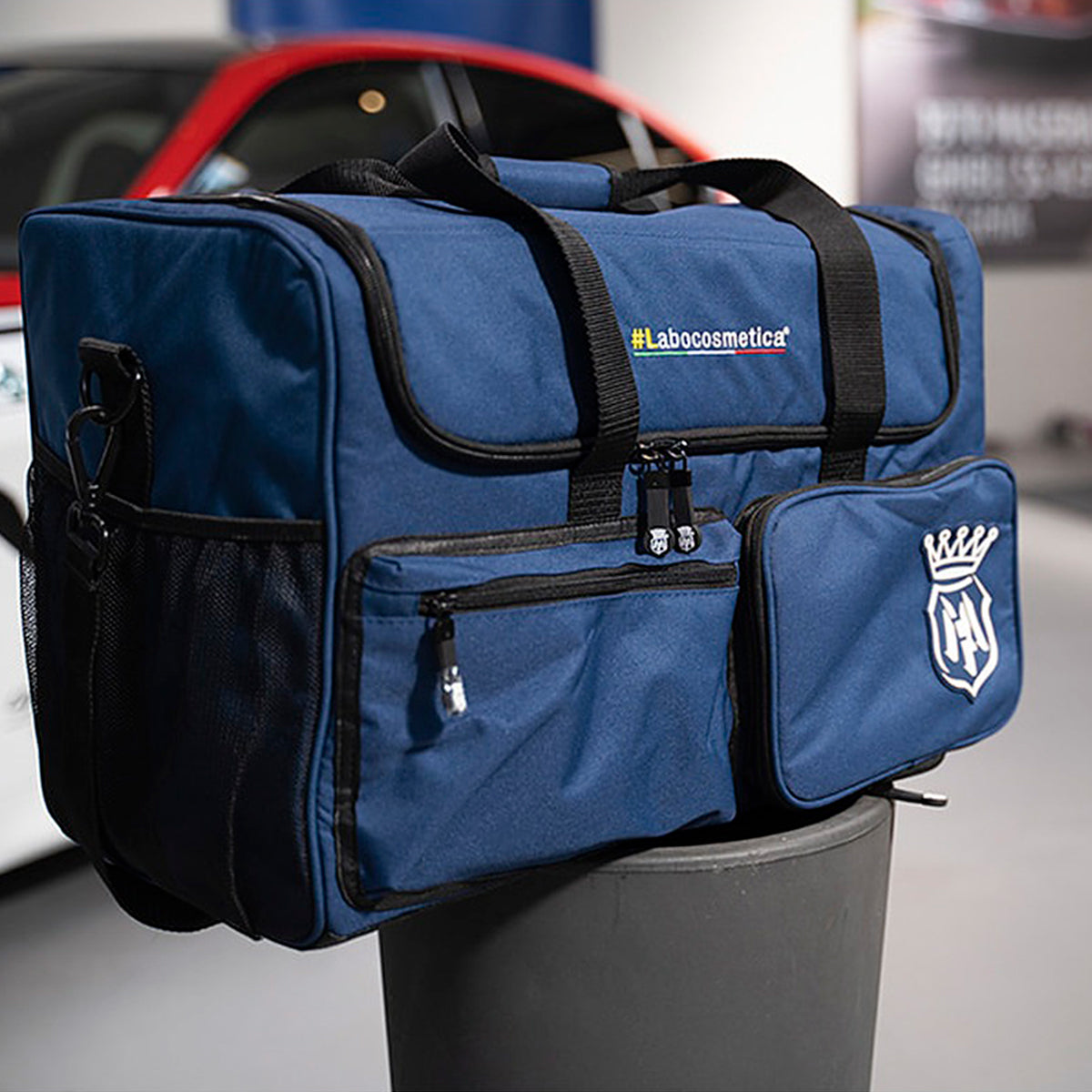 A blue cosmetic travel duffel bag with black handles and zippers, featuring a white crown-and-letter logo on the front pocket and a small 'Labooocosmetic' label, displayed on a gray cylindrical stand with a blurred car in the background.