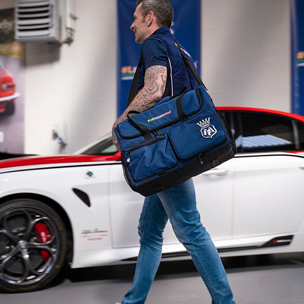 A man carrying a large blue and black Fingertip duffel bag over his shoulder, walking past a white sports car in a garage setting.