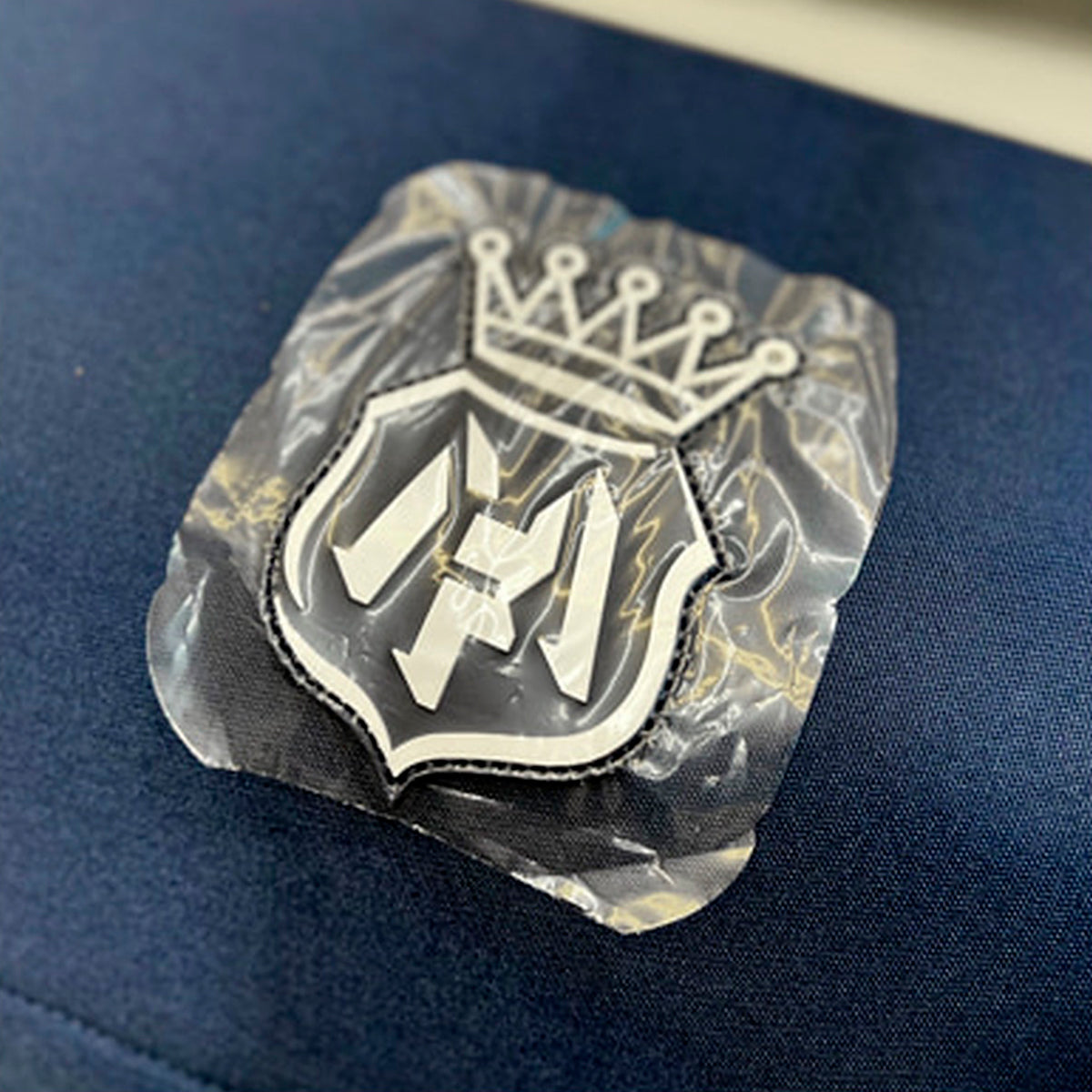A close-up of a custom embroidered patch featuring a white crown and shield design with the letters 'HF', sealed in clear plastic packaging and placed on a dark blue textured fabric background.