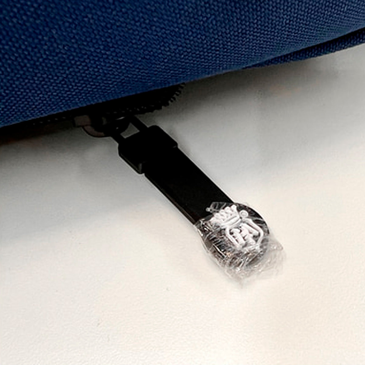Close-up of a zipper pull on a blue fabric item, with the pull wrapped in clear plastic on a white surface.