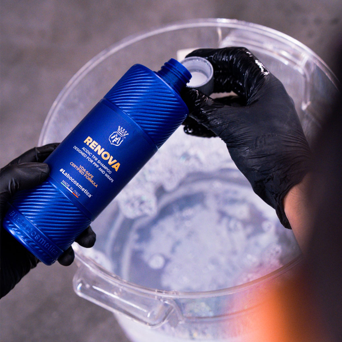 Person wearing black gloves pouring a blue bottle labeled 'Renova' into a clear plastic container filled with water.